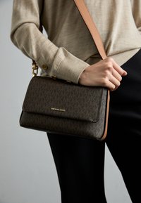 Brown patterned handbag with a gold logo, featuring a flap closure, leather accents, and a detachable shoulder strap, held in a hand.
