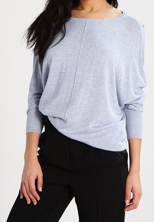 Maglione azzurro chiaro, dalla vestibilità ampia con scollatura larga e maniche a costine. Texture morbida e dettagli di cucitura evidenti lungo il davanti. Abbinato a pantaloni neri.