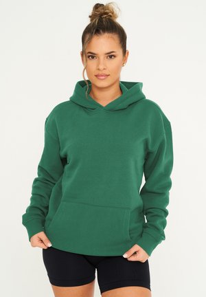 Green cotton hoodie with a front pocket, drawstring hood, and relaxed fit. Long sleeves with cuffed edges, paired with black shorts.