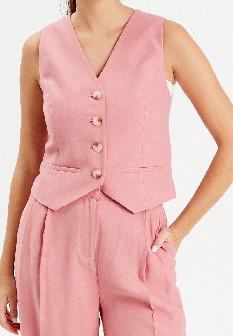 Gilet rose sans manches boutonné avec ourlet en encoche porté avec un pantalon taille haute assorti, une main dans la poche, sur fond blanc.