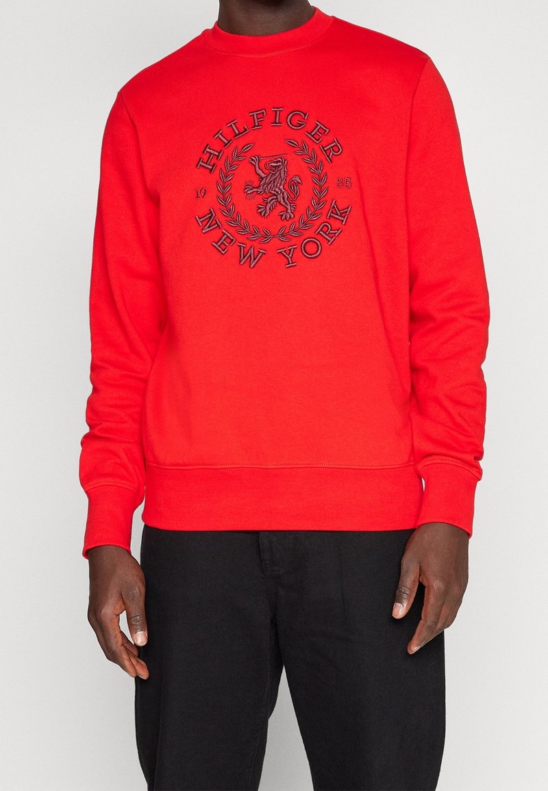 Personne portant un sweat-shirt rouge vif avec le texte brodé en noir « Hilfiger New York 1985 » et un blason, associé à un pantalon noir.