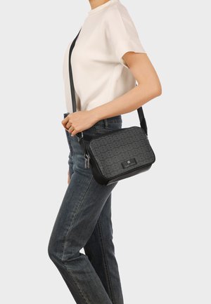 Donna che indossa una camicia bianca e jeans porta una borsa a tracolla nera con motivo e tracolla regolabile sul lato destro.