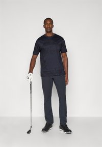Námornícka košeľa s krátkym rukávom má jemný mriežkovaný vzor, kombinovaná s tmavými nohavicami a čiernymi golfovými topánkami. Biela golfová rukavica je držaná v ľavej ruke.