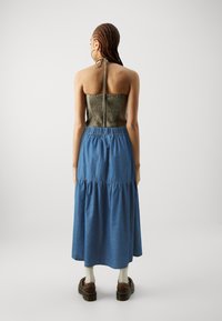 Cheveux tressés avec un haut vert olive de style harnais, mettant en valeur un corsage ajusté et une jupe en denim bleu à volants. Chaussures marron et chaussettes blanches visibles.