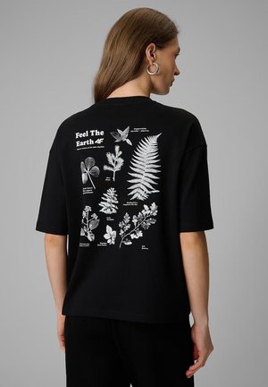 Kobieta z długimi włosami ma na sobie czarną koszulkę z białymi botanicznymi ilustracjami i napisem "Feel The Earth 4F" na plecach.