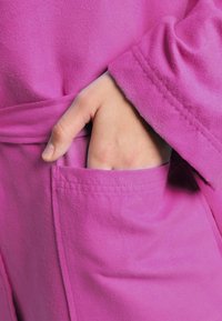 Accappatoio in pile rosa acceso con cintura, tasca laterale e maniche arrotolate. La texture appare morbida e confortevole.