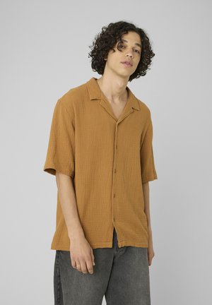 Jeune homme aux cheveux bouclés portant une chemise moutarde ample à manches courtes et un pantalon gris, debout devant un fond gris.