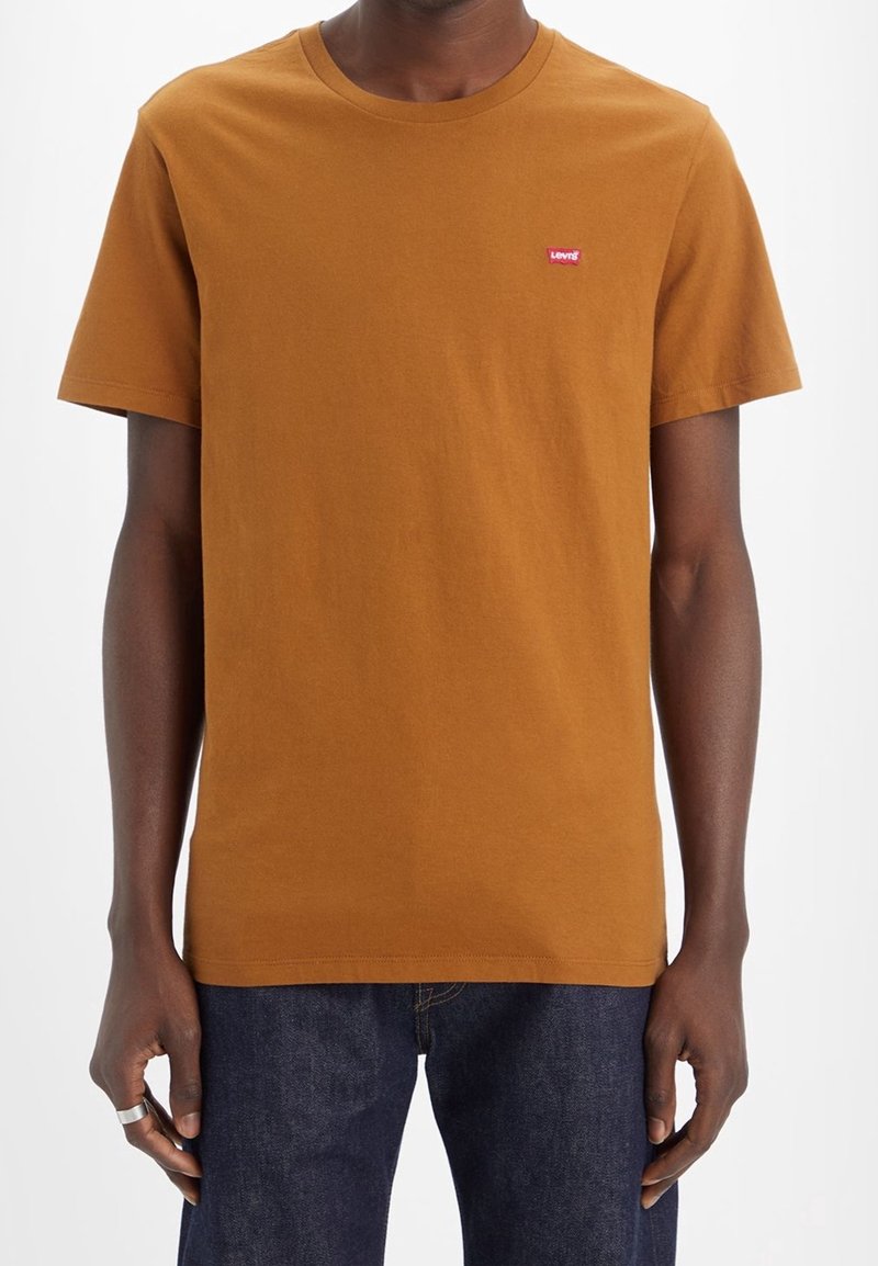 Homme portant un t-shirt marron uni à manches courtes avec un petit logo rouge Levi's sur la poitrine et un jean bleu foncé.