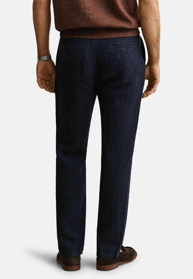 Mann trägt dunkelblaue Nadelstreifenhose, braunes Hemd, braune Loafers und silberne Armbanduhr, steht mit dem Rücken zur Kamera vor weißem Hintergrund.