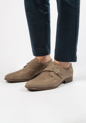 Chaussures en suede marron avec un bout pointu, une sangle à l'avant et un détail de boucle en argent. Semelle plate marron, portées avec un pantalon sombre.