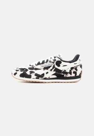 Baskets Reebok avec motif imprimé vache noir et blanc, lacets blancs et semelle beige sur fond gris clair uni.