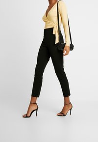 Gelbe Wickelbluse mit langen Ärmeln, schwarze, figurbetonte Hose mit Knöchelschlitzen und schwarze Riemchensandalen. Kleine schwarze Umhängetasche.