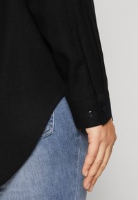 Black button-up shirt with long sleeves, textured fabric, and a curved hem. Features black buttons at the cuff and worn with light blue jeans.