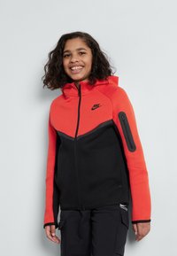 Nike zip-up hette i klar orange og svart, med et kontrasterende øverste design, glidelås-lomme på siden og teksturert materiale.