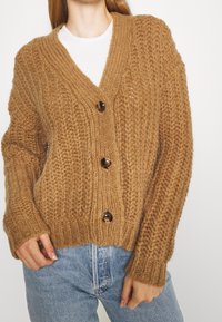 Brauner Strickcardigan mit V-Ausschnitt, ausgestattet mit drei großen dunklen Knöpfen, gerippten Bündchen und lockerer Passform. Strukturmuster im gesamten Kleidungsstück.