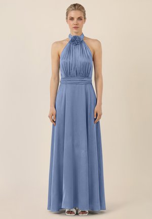 Vrouw draagt een vloerlang halterjurk in leisteengrijs met plooien en een roosdetail bij de halslijn, staand tegen een effen achtergrond.