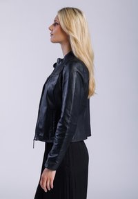 Černá kožená bunda s přiléhavým designem, zapínáním na zip a límcem. Hladký povrch a zkrácená délka. Modelka má pod ní černé šaty.