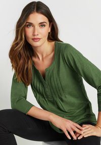 Groene lange mouwtop met een ronde halslijn, voorzien van verticale plooien, knoopdetails en een zachte, gestructureerde stof.