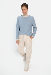 Lichtblauwe langesleeve trui over een beige coltrui, gecombineerd met losse beige broek en lichtgrijze sneakers.