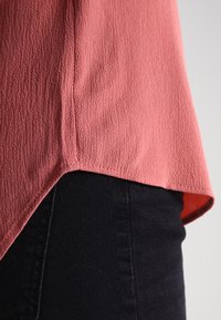 Koralfarbenes, strukturiertes Oberteil mit einem Saum, der an den Seiten tiefer gezogen ist, kombiniert mit schwarzem Denim. Der Stoff scheint leicht und leicht zerknittert zu sein.