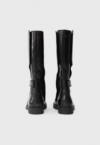 Botine din piele neagră, până la genunchi, cu o textură netedă, având un detaliu cu bandă și tocul jos. Vedere din spate care arată cusăturile laterale.