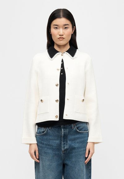 Γυναίκα με λευκό σακάκι με κουμπιά πάνω από μαύρο τοπ με γιακά και μπλε τζιν, που στέκεται μπροστά σε απλό φωτεινό φόντο.