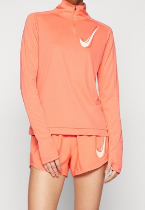 Person iført koralrød langærmet sportstop med kvart lynlås og matchende shorts, begge med store hvide Nike Swoosh-logoer.
