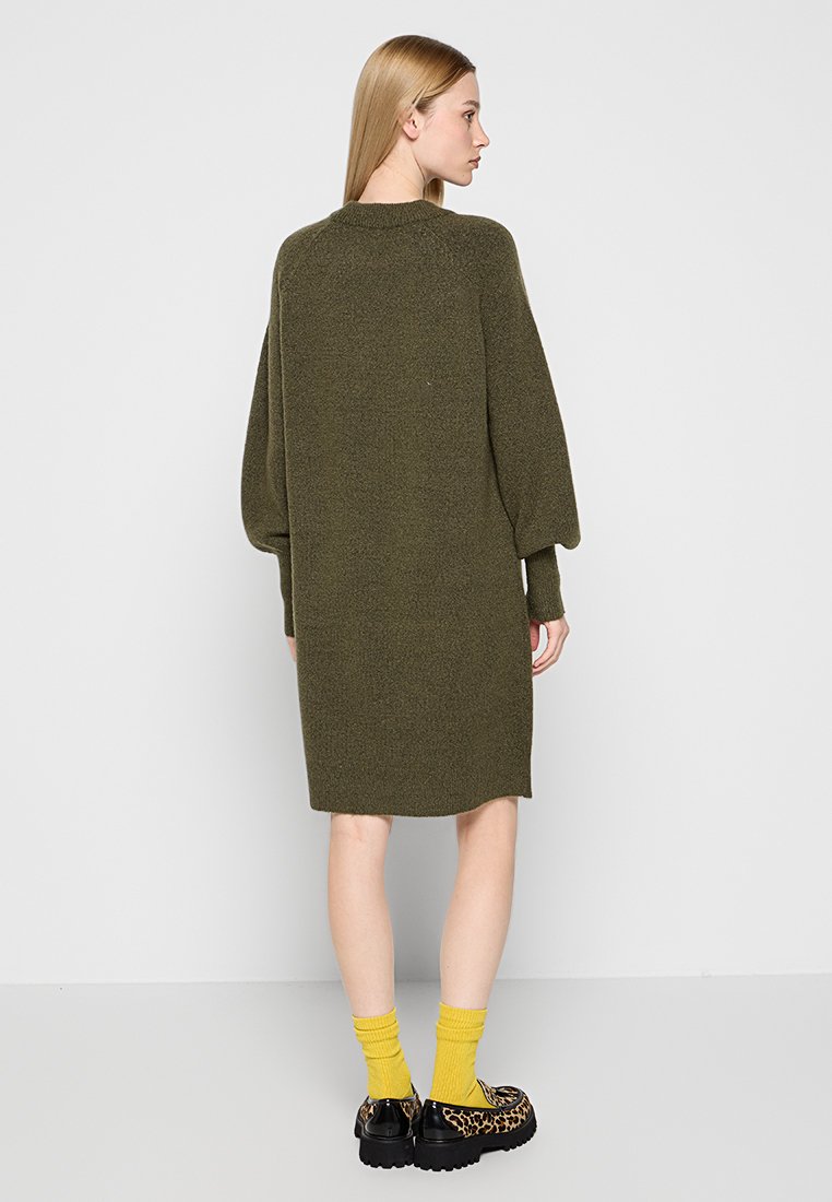 Langes, übergroßes olivgrünes Strickkleid mit Ballonärmeln, weichem Material und geripptem Saum. Kombiniert mit gelben Socken und Schuhen im schwarzen Leopardenmuster.