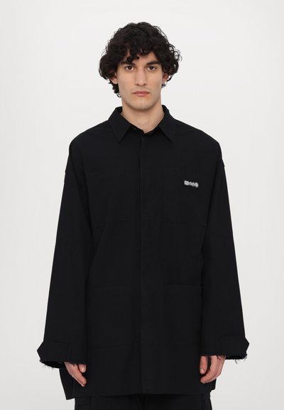 Camisa de botón negra de gran tamaño con mangas largas, dos bolsillos frontales, dobladillo deshilachado y un pequeño parche con logo en el pecho. Tejido de algodón.