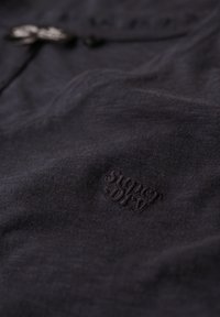 Tela de algodón negra con una textura suave, que presenta un logo de 'Superdry' embosado en una esquina. Diseño minimalista con sutiles variaciones de color.