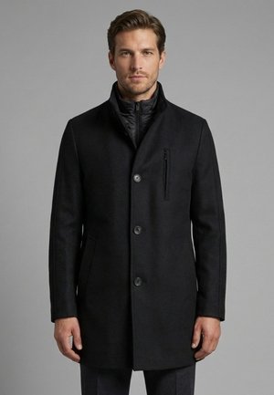 Mann trägt einen schwarzen, geknöpften Wollmantel über einer gesteppte Reißverschlussjacke und steht vor einem schlichten grauen Hintergrund.