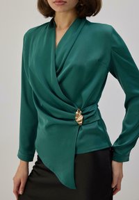 Mujer vistiendo una blusa cruzada de satén color teal con mangas largas, adornada con un broche dorado en forma de hoja en la cintura, combinada con una falda negra.