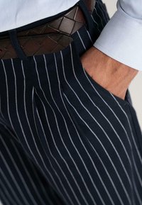 Man in marineblauwe streepbroek en een geweven bruine leren riem, met de rechterhand casual in de zak.