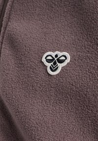 Lila fleece-stoff med en vit broderad bi-logotyp med svarta detaljer, placerad på vänster sida av plagget.