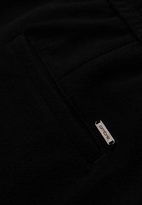 Gros plan sur un tissu noir avec une petite étiquette en métal portant l'inscription "ombre" cousue près d'une couture ou du bord d'une poche.