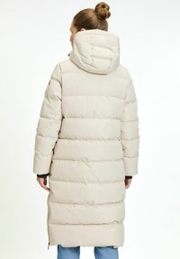 Vrouw met een knot in haar haar, gekleed in een lange, crèmekleurige pufferjas met capuchon, staande met de rug naar de camera tegen een eenvoudige witte achtergrond.