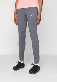Grå treningsleggings med en rosa Nike-logo, med slank passform, glatt tekstur og kontrasterende sømmer. Brukt med svarte fotballsko.