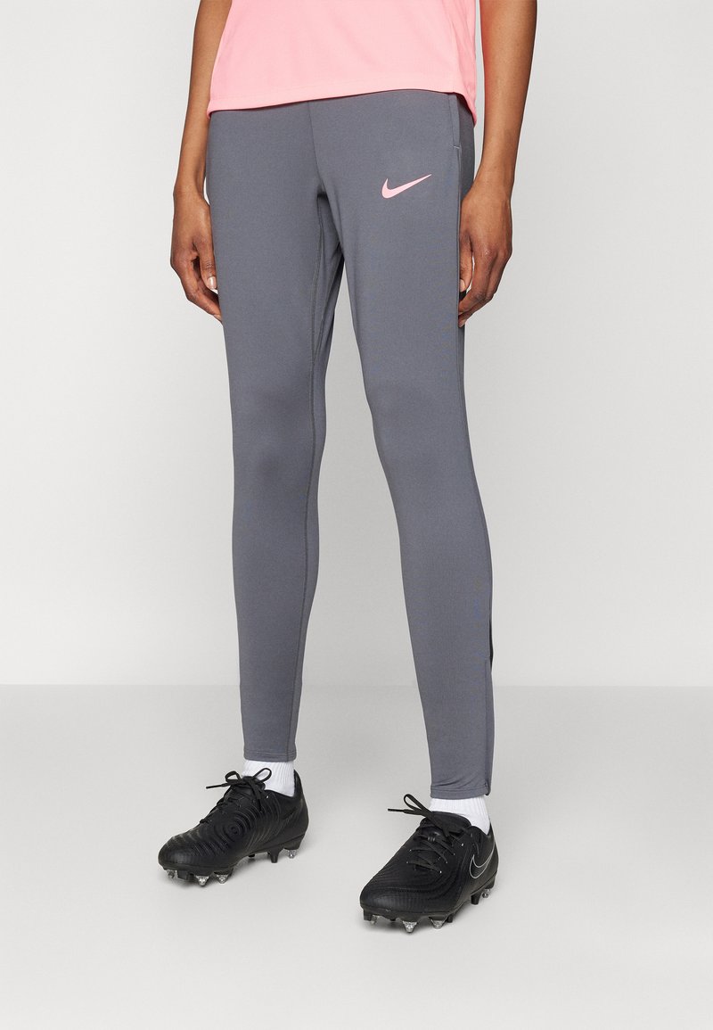 Grå treningsleggings med en rosa Nike-logo, med slank passform, glatt tekstur og kontrasterende sømmer. Brukt med svarte fotballsko.