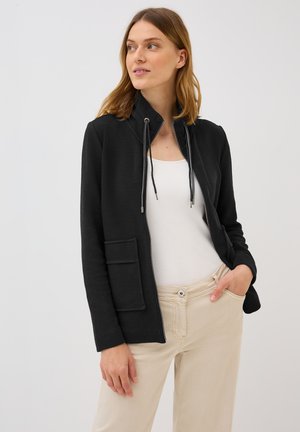 Eine Frau lächelt, während sie eine schwarze Zip-Jacke über einem weißen Oberteil trägt, mit ihren Händen in den Taschen ihrer beigen Hose.