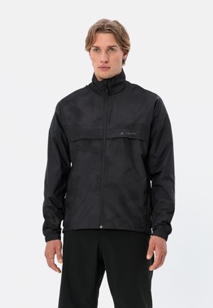 Mann trägt eine schwarze Jacke mit Reißverschluss und hohem Kragen sowie einem dezenten Camouflagemuster, steht vor einem schlichten hellen Hintergrund.