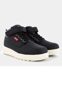 Stivali neri alti fino alla caviglia con una texture liscia, dotati di una fascia in Velcro e occhielli dorati, montati su una robusta suola bianca. Dettaglio logo rosso presente.