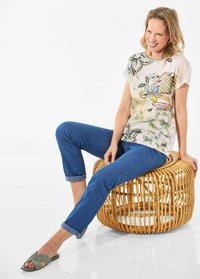 Bloemmotieven korte mouwen top in lichte kleuren, gecombineerd met blauwe jeans met opgerolde mouwen en olijfgroene platte sandalen, zittend op een geweven kruk met dikke rand.