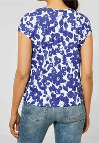 Žena v bílém tričku s krátkým rukávem s modrým květinovým vzorem a světle modrých džínách, viděná zezadu.