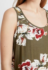 Débardeur vert olive avec de grands motifs floraux blancs et rouges, avec un décolleté rond et de larges bretelles. Texture de tissu lisse.