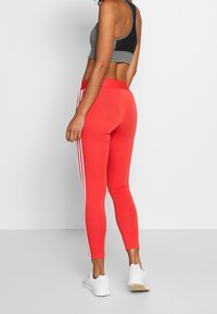 Czerwone legginsy z białymi paskami po bokach, wykonane z elastycznego materiału, w połączeniu z szarym biustonoszem sportowym, zaprojektowane do aktywności fizycznej.