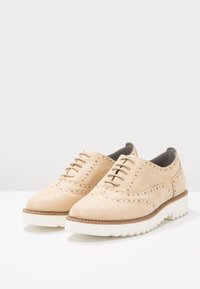 Beige semišové boty s broušeným designem, kulatou špičkou, tkaničkami a silnou bílou gumovou podrážkou s texturovaným gripem.