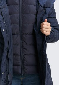 Granatowa kurtka puchowa z zamkiem z przodu, pikowanym wzorem i kapturem. Materiał wydaje się lekki i wodoodporny, z zapięciami na napy.