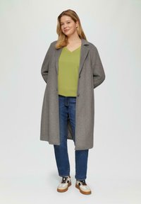 Vrouw staand in een lange grijze jas, lichtgroen V-hals top, blauwe spijkerbroek en witte sneakers met zwarte accenten.