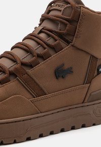 Brązowe wysokie trampki wykonane z teksturowanego materiału z kontrastującymi sznurówkami i czarnym akcentem logo. Posiadają solidną podeszwę zapewniającą przyczepność.