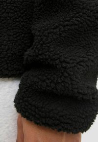 Manica di giacca in pile nera con una superficie testurizzata e morbida. Presenta un orlo leggermente arrotolato, che mette in evidenza un tessuto spesso e morbido.